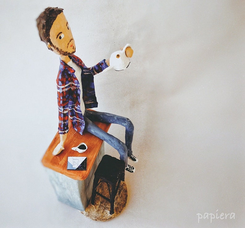 May include: A handmade figurine of a person sitting on a wooden stool with a cup of coffee and a book. The person is wearing a plaid shirt and jeans. The figurine is made of paper mache and painted in a realistic style.