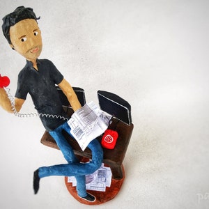 May include: A figurine of a person sitting at a desk with a red telephone, a computer, and papers. The figurine is made of papier-m&acirc;ch&eacute; and is painted in realistic colours.