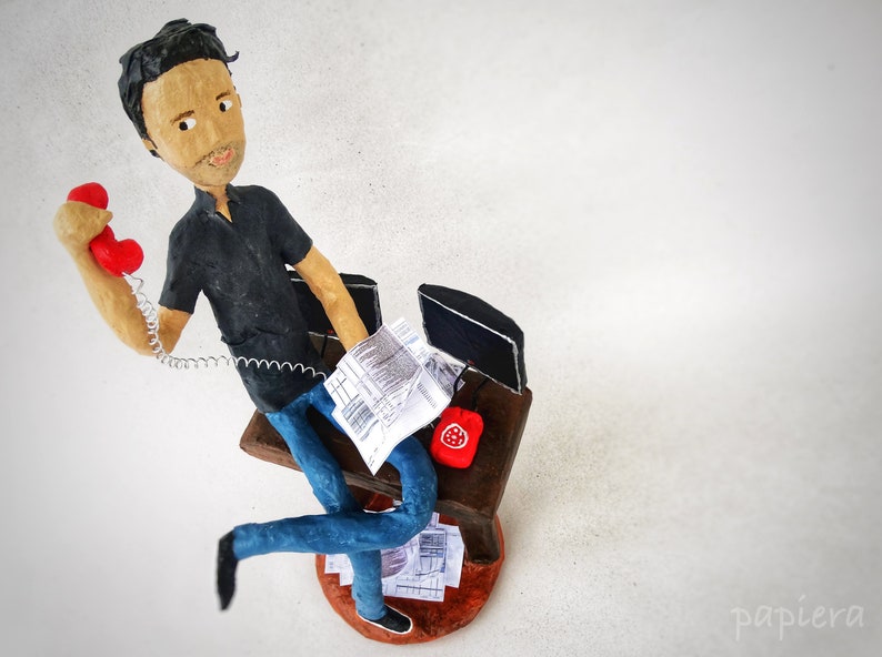 May include: A figurine of a person sitting at a desk with a red telephone, a computer, and papers. The figurine is made of papier-m&acirc;ch&eacute; and is painted in realistic colours.