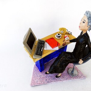 May include: A figurine of a woman sitting at a desk with a computer, a phone, and a cup of coffee. The desk is made of wood and has a blue and white paint job. The woman is wearing a black dress and has grey hair.
