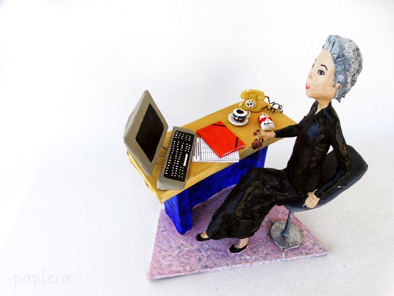 May include: A figurine of a woman sitting at a desk with a computer, a phone, and a cup of coffee. The desk is made of wood and has a blue and white paint job. The woman is wearing a black dress and has grey hair.