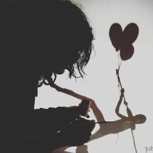 May include: A person's silhouette creating a shadow puppet of a figure holding two heart-shaped balloons on a white wall. The person is holding a paintbrush and a small bottle of paint.