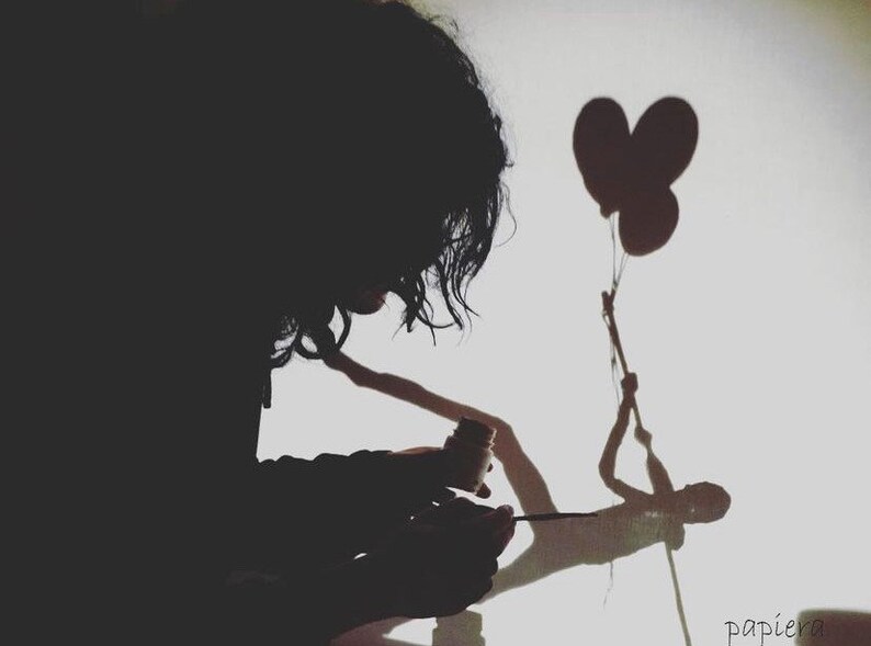 May include: A person's silhouette creating a shadow puppet of a figure holding two heart-shaped balloons on a white wall. The person is holding a paintbrush and a small bottle of paint.