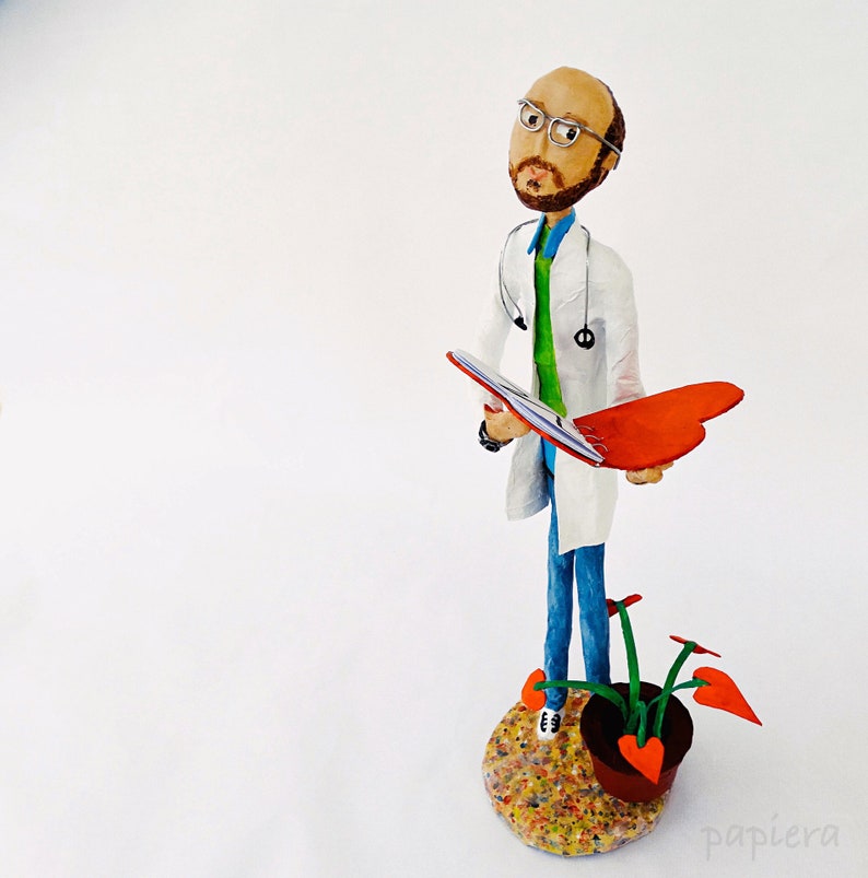 May include: A handmade figurine of a doctor wearing a white coat, glasses, and a stethoscope. The doctor is holding a red folder and standing on a colourful base with a potted plant.