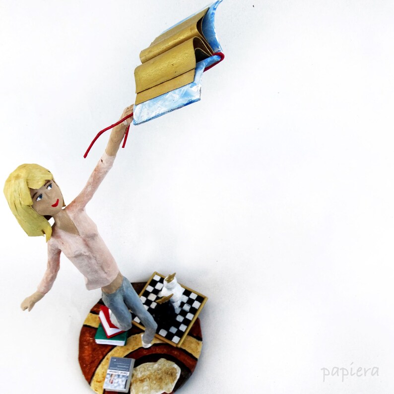 May include: A figurine of a woman with blonde hair holding a stack of books in her hand. The figurine is standing on a base with a chessboard, books, and a rug.