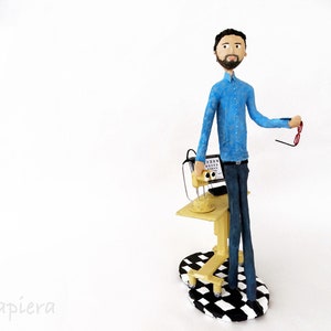 May include: A figurine of an optometrist wearing a blue shirt and holding a pair of glasses. The figurine is standing next to a miniature eye exam machine with a black and white chequered base.