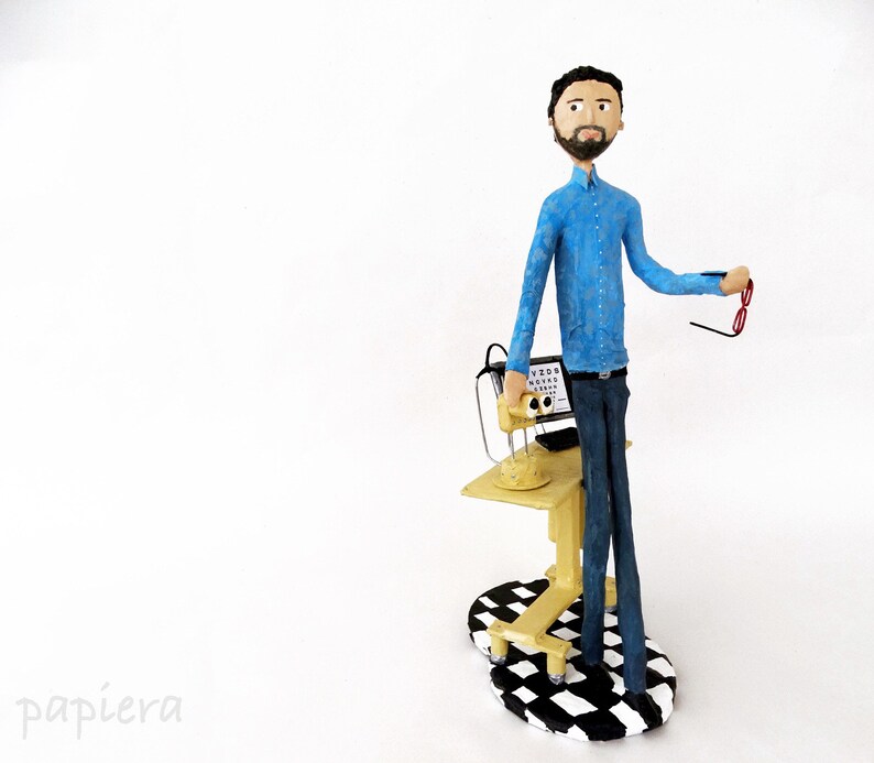May include: A figurine of an optometrist wearing a blue shirt and holding a pair of glasses. The figurine is standing next to a miniature eye exam machine with a black and white chequered base.