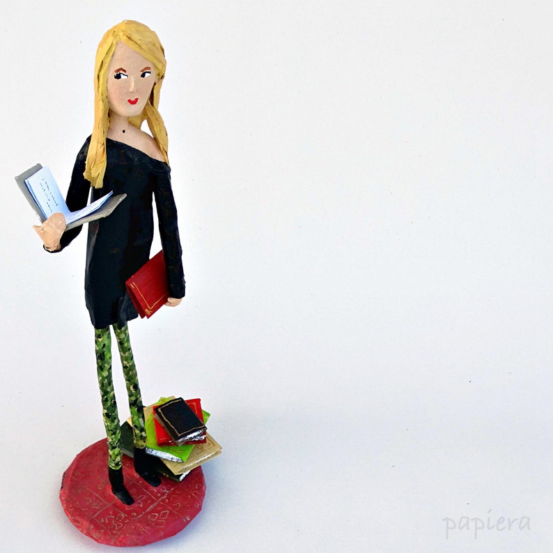 May include: A paper mache figurine of a woman with blonde hair wearing a black dress and green leggings. She is holding a book and standing on a red base with a stack of books.