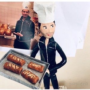 May include: A paper mache figurine of a chef wearing a white chef's hat and black jacket, holding a baking tray with four loaves of bread. The figurine is standing in front of a photo of a person in a chef's hat and jacket holding a baking tray with loaves of bread.