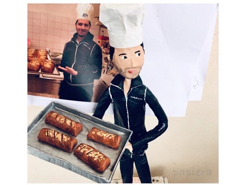 May include: A paper mache figurine of a chef wearing a white chef's hat and black jacket, holding a baking tray with four loaves of bread. The figurine is standing in front of a photo of a person in a chef's hat and jacket holding a baking tray with loaves of bread.