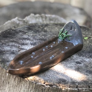May include: A brown ceramic incense burner shaped like a wooden plank with a small tombstone at the end. The tombstone is gray and has the letters "RIP" on it. There is a green plant growing out of the top of the tombstone.