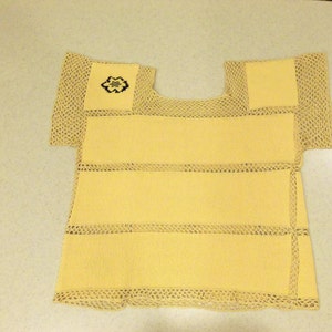 May include: A yellow knitted top with a square neckline and short sleeves. The top has horizontal bands and is trimmed with beige, open-weave crochet. A small embroidered design is on the upper left.