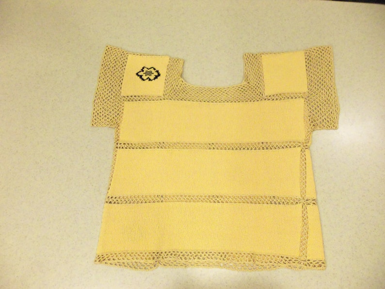 May include: A yellow knitted top with a square neckline and short sleeves. The top has horizontal bands and is trimmed with beige, open-weave crochet. A small embroidered design is on the upper left.