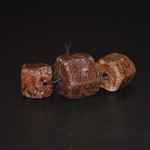 Könnte beinhalten: Drei quadratische, dunkelrote Edelsteinkugeln, die auf einer schwarzen Schnur aufgefädelt sind. Die Kugeln haben eine raue, strukturierte Oberfläche.