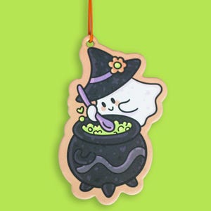 May include: A hanging ornament featuring a white ghost wearing a black witch hat with a purple band and a small orange flower. The ghost is stirring a black cauldron filled with a glowing green liquid using a purple spoon, suspended by a red string.