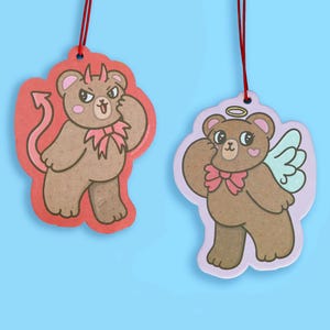 May include: Two hanging ornaments featuring brown bears. The left bear has red devil horns and a tail on a red background. The right bear has a yellow halo and light blue wings on a light purple background. Both bears wear pink bows.
