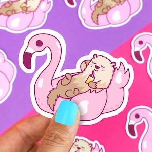 May include: A pink flamingo pool float with a brown and white otter relaxing inside. The otter is holding a yellow drink and has a relaxed expression.
