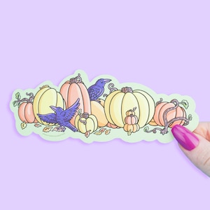 May include: A sticker featuring a collection of pumpkins in various sizes and colors, including yellow, orange, and peach. Two purple crows are perched among the pumpkins. The sticker has a light green border and is held by a hand with a purple painted nail.