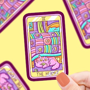 May include: A tarot card illustration of The Hermit, featuring a pink cat hiding under a colorful patchwork quilt. The card is pink and purple with the Roman numeral IX and the text "THE HERMIT" at the bottom.