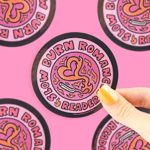 May include: Round sticker with a black border and a pink background. The sticker features the words "BURN ROMANCE SLOW READER" in a circular pattern. The center design includes a heart and flames in orange and pink.