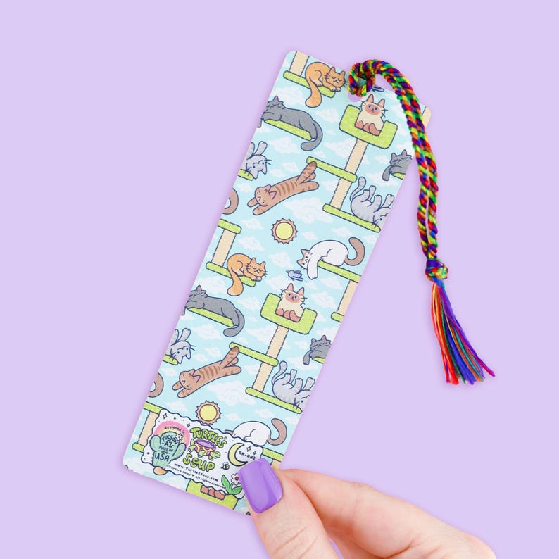 May include: A rectangular bookmark featuring a repeating pattern of cartoon cats on cat trees against a light blue sky background. The bookmark has a rainbow-colored tassel attached to the top. The text "Turtles Soup" is visible at the bottom.
