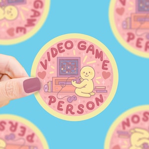 May include: A round sticker with a light pink background and a yellow border. The sticker features a cartoon person playing a video game. The words "VIDEO GAME PERSON" are written around the edge of the sticker. Hearts and stars are scattered around the design.