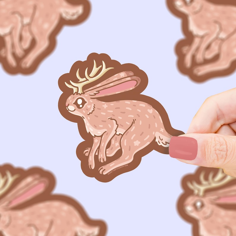 Puede incluir: Una pegatina de un jackalope de dibujos animados, criatura m&iacute;tica con cuernos y orejas rosas. El jackalope es de color marr&oacute;n claro con detalles blancos, sobre un fondo lila claro.