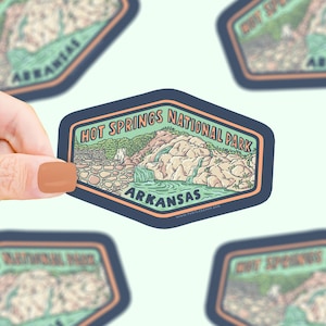 May include: A sticker featuring a scenic illustration of Hot Springs National Park in Arkansas. The design includes a waterfall, rocks, and the park's name in a vintage style. The sticker has a navy blue border with an orange outline.