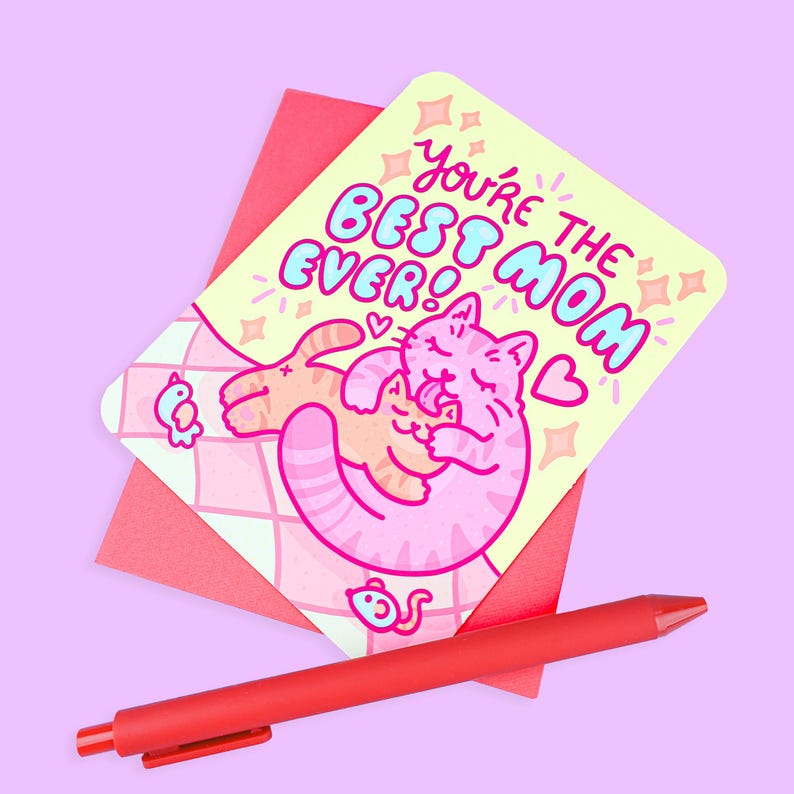 May include: A greeting card with the text "You're the BEST MOM EVER!" features a cartoon cat hugging another cat. The card is on top of a red envelope, with a red pen in the foreground. The background is a light purple color.