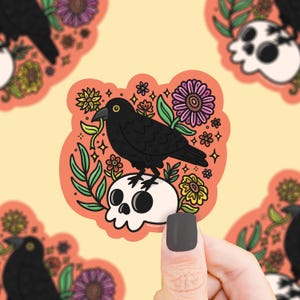 May include: A sticker featuring a black crow perched on a white skull, surrounded by colorful flowers and green leaves. The sticker has an orange background and is a whimsical design.