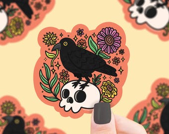 May include: A sticker depicting a black crow atop a white skull, encircled by vibrant flowers and green leaves. The sticker is set against an orange backdrop, showcasing a whimsical design.