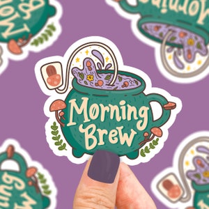 May include: A green cauldron with a purple liquid inside and a white steam coming out of the top. The cauldron has a handle and is decorated with mushrooms and leaves. The text "Morning Brew" is written in white on the cauldron.