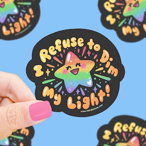 May include: A black sticker with a rainbow star illustration and the text "Refuse to Dim My Light!". The star has a smiling face and is surrounded by colorful rays and sparkles. The sticker is held by a hand with a pink nail polish.