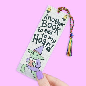 May include: A whimsical bookmark featuring a cartoon goblin holding a book, with the text "Another BOOK to add to my HOARD." The bookmark has a colorful tassel and is set against a brick wall background.