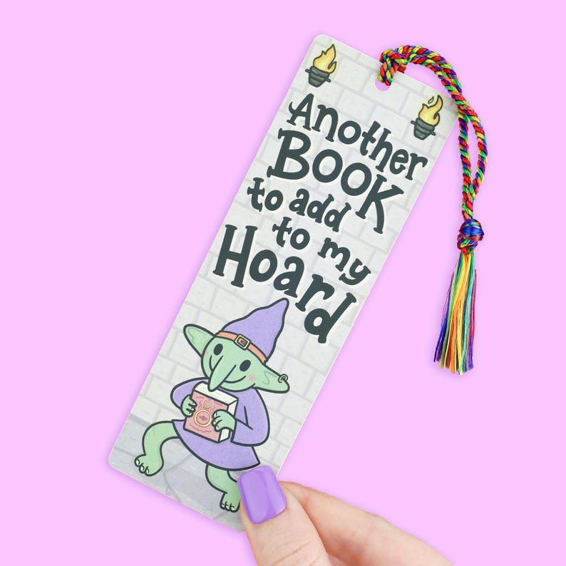 May include: A whimsical bookmark featuring a cartoon goblin holding a book, with the text "Another BOOK to add to my HOARD." The bookmark has a colorful tassel and is set against a brick wall background.