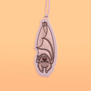 May include: A hanging bat-shaped ornament with a smiling face and outstretched wings. The bat is light gray with darker gray details and a purple string for hanging. The background is a solid orange color.