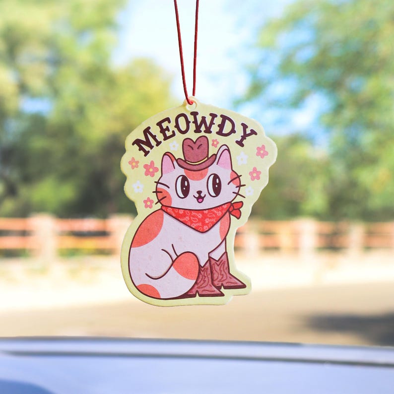 May include: A yellow car air freshener with a cartoon cat wearing a cowboy hat and bandana. The text "MEOWDY" is written above the cat.