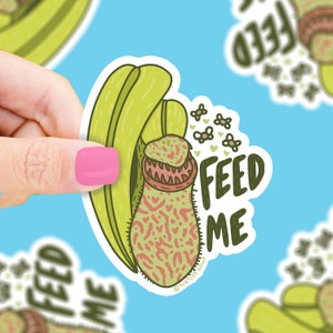 May include: A white sticker featuring a green pitcher plant with a red and pink mouth. The plant is surrounded by small pink flowers and flies. The text "FEED ME" is written in black on the sticker.