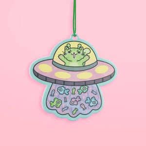 May include: A whimsical air freshener featuring a cartoon cat in a UFO. The UFO is light blue, pink, and yellow, with a green string for hanging. The UFO is emitting a beam of cartoon fish. The background is pink.