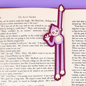 Puede incluir: Un marcap&aacute;ginas de mono morado con un patr&oacute;n de lunares rosas y una cara blanca. El marcap&aacute;ginas est&aacute; colocado en una p&aacute;gina de un libro con el texto "The Secret Garden".