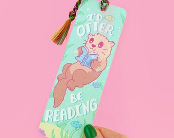 Puede incluir: Un marcapáginas verde y azul con una nutria de dibujos animados leyendo un libro. El marcapáginas tiene el texto "I'd otter be reading".