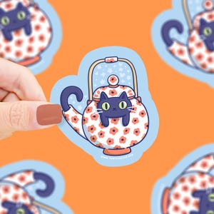 May include: A sticker featuring a cartoon black cat peeking out of a teapot. The teapot is white with a pattern of orange flowers and a blue handle. The background is orange.