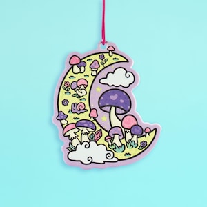 May include: A whimsical, crescent moon-shaped decoration with a pink border and a pink string for hanging. The design features colorful mushrooms, flowers, clouds, and a snail. The color palette includes shades of purple, pink, yellow, and white.