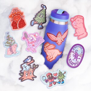 Puede incluir: Una colecci&oacute;n de pegatinas coloridas con ilustraciones caprichosas, que incluyen un oso envuelto en una manta, un &aacute;rbol con una cara y un perro rosa bebiendo t&eacute;. Una botella de agua azul est&aacute; decorada con pegatinas de un b&uacute;ho y un conejo.