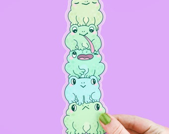 May include: A stack of five frog stickers in various shades of green and blue. The frogs are all smiling and have different expressions. The stickers are arranged in a vertical stack, with the largest frog at the bottom and the smallest frog at the top. The stickers are on a purple background. The text "www.Turtlesoup.com" is at the bottom of the sticker.