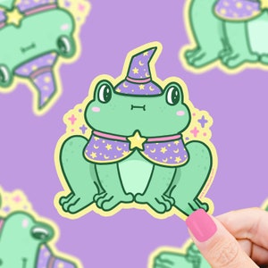 May include: A green frog wearing a purple and yellow wizard hat and a purple and yellow robe with stars. The frog has a big smile and is looking at the viewer.