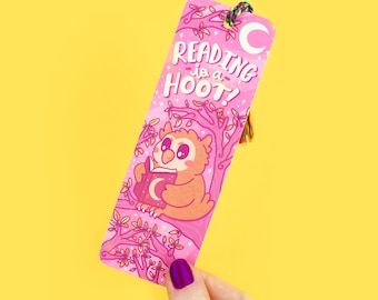 Puede incluir: Marcapáginas rosa con un búho de dibujos animados leyendo un libro. El marcapáginas tiene el texto "Reading is a Hoot!" y una luna creciente.