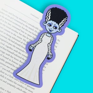 May include: A die-cut bookmark featuring a stylized illustration of the Bride of Frankenstein with tall black hair, white streaks, pale blue skin, and a long white gown. The character has visible stitches on the neck and arms, set against a light purple border.