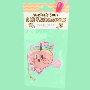May include: A peach-shaped air freshener in a clear plastic package. The air freshener has a cartoon peach design with a surprised expression and a cloud of vapor. The packaging reads "Turtle's Soup Air Freshener" and "Peach Toot".