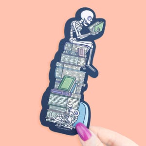 May include: A sticker depicting a skeleton reading a book while seated on a pile of books. The skeleton is white, and the books are various shades of green, purple, and white. A skeleton dog is at the base.